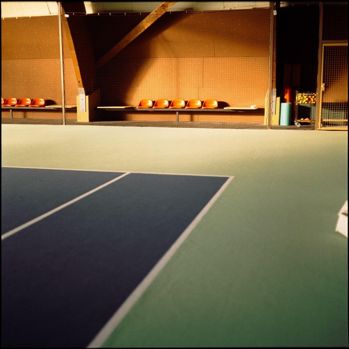 A tennis court with orange seats along the back wall is illuminated by sunlight.