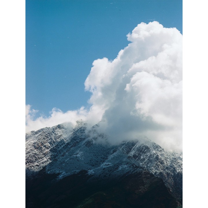 A large cloud covers the peak of a snow-dusted mountain under a blue sky.