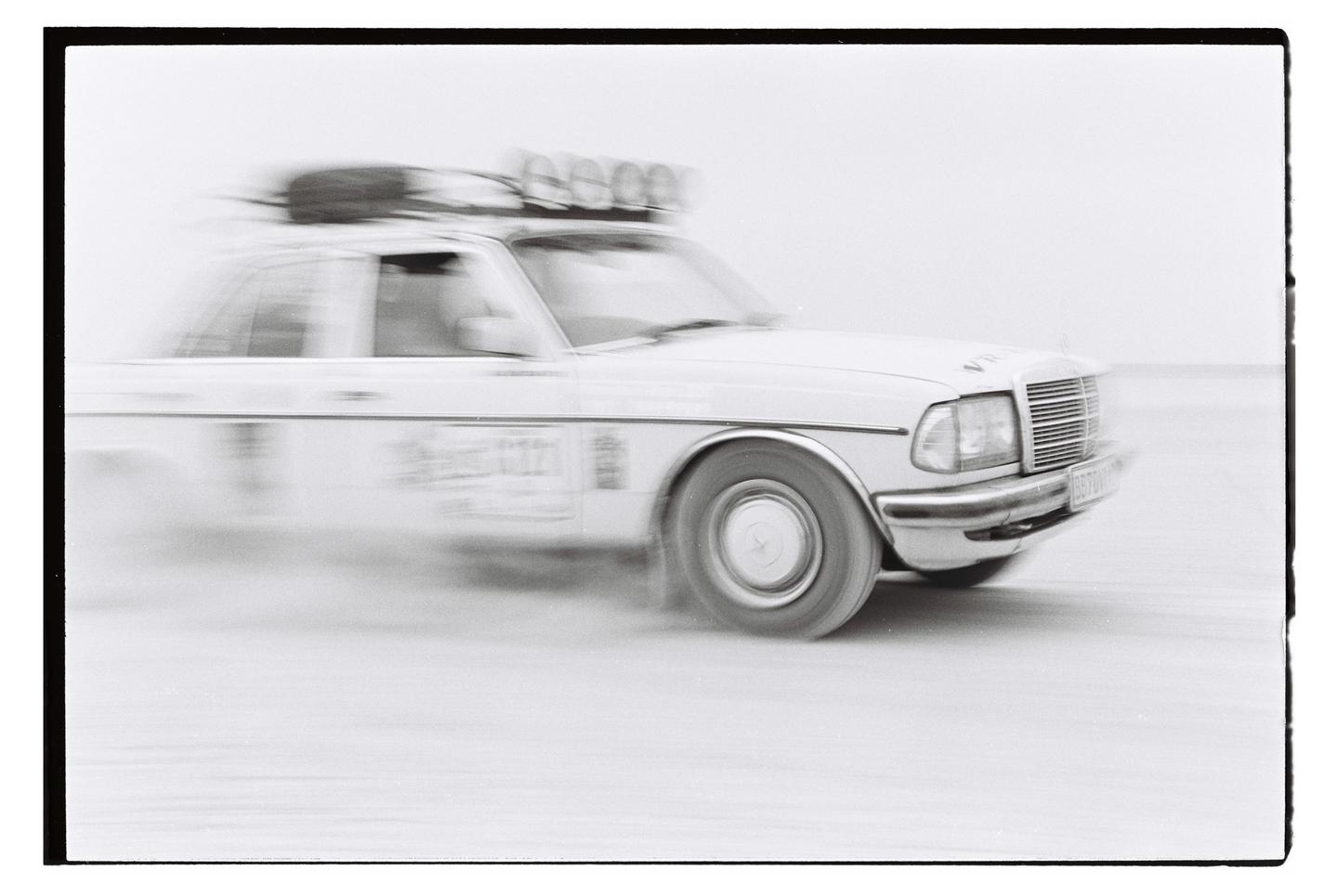 A white car with a roof rack is captured in motion, blurring the background.