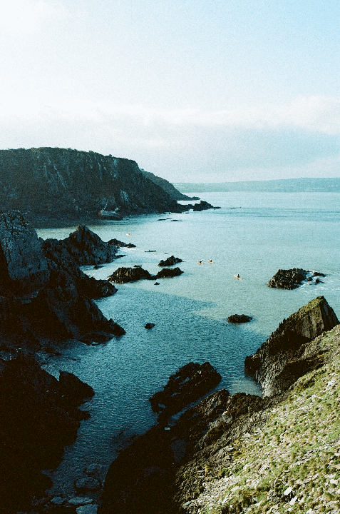 A rugged coastline with dark cliffs and rocky outcrops extends into the sea, where several small boats are visible on the water.