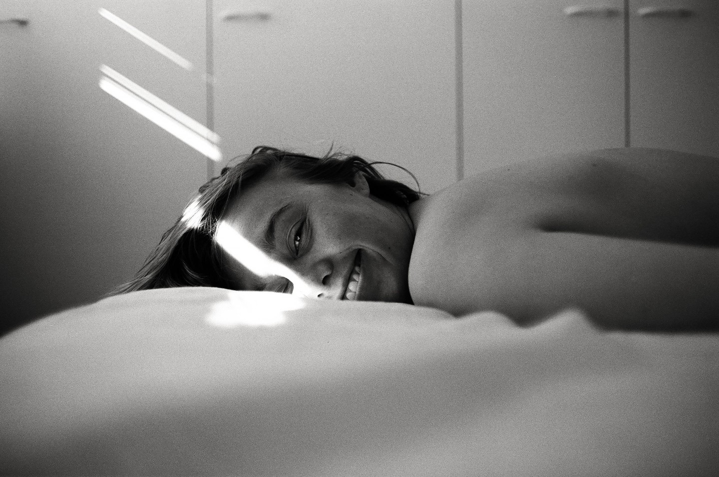 A woman lies on a bed, smiling at the camera with a beam of light across her face.