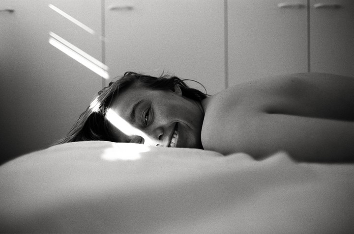 A woman lies on a bed, smiling at the camera with a beam of light across her face.