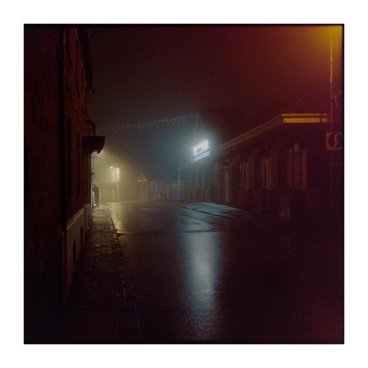 A street at night is illuminated by a bright light and a streetlamp, with a person standing in the distance.