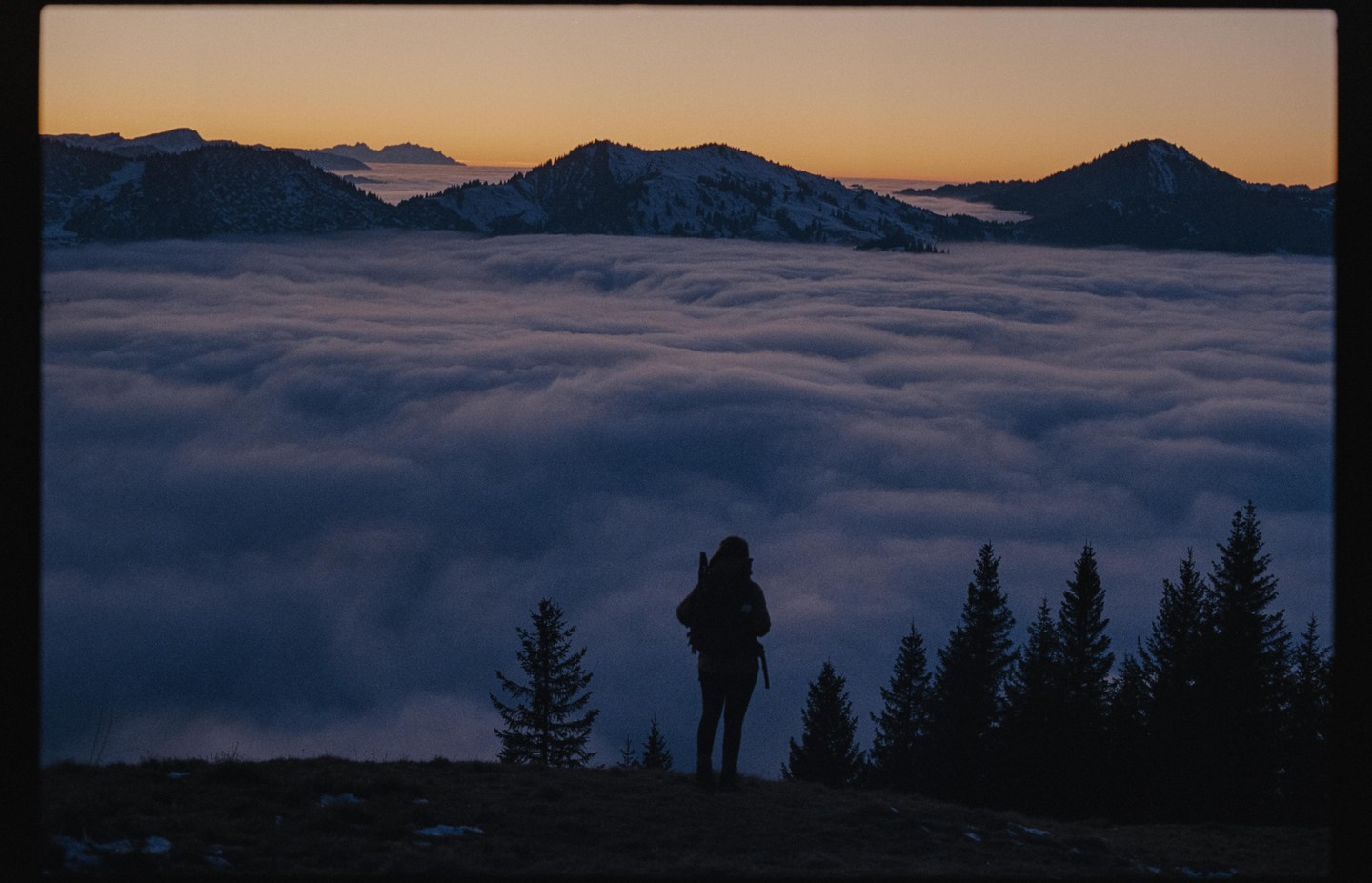 A person stands on a hill overlooking a valley filled with clouds at sunset.