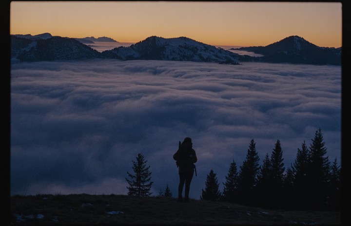 A person stands on a hill overlooking a valley filled with clouds at sunset.