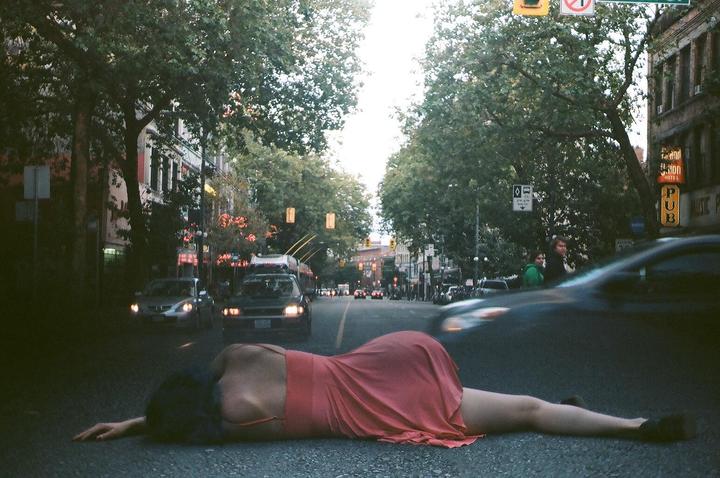 A woman in a red dress is lying on a city street with cars and trees in the background.