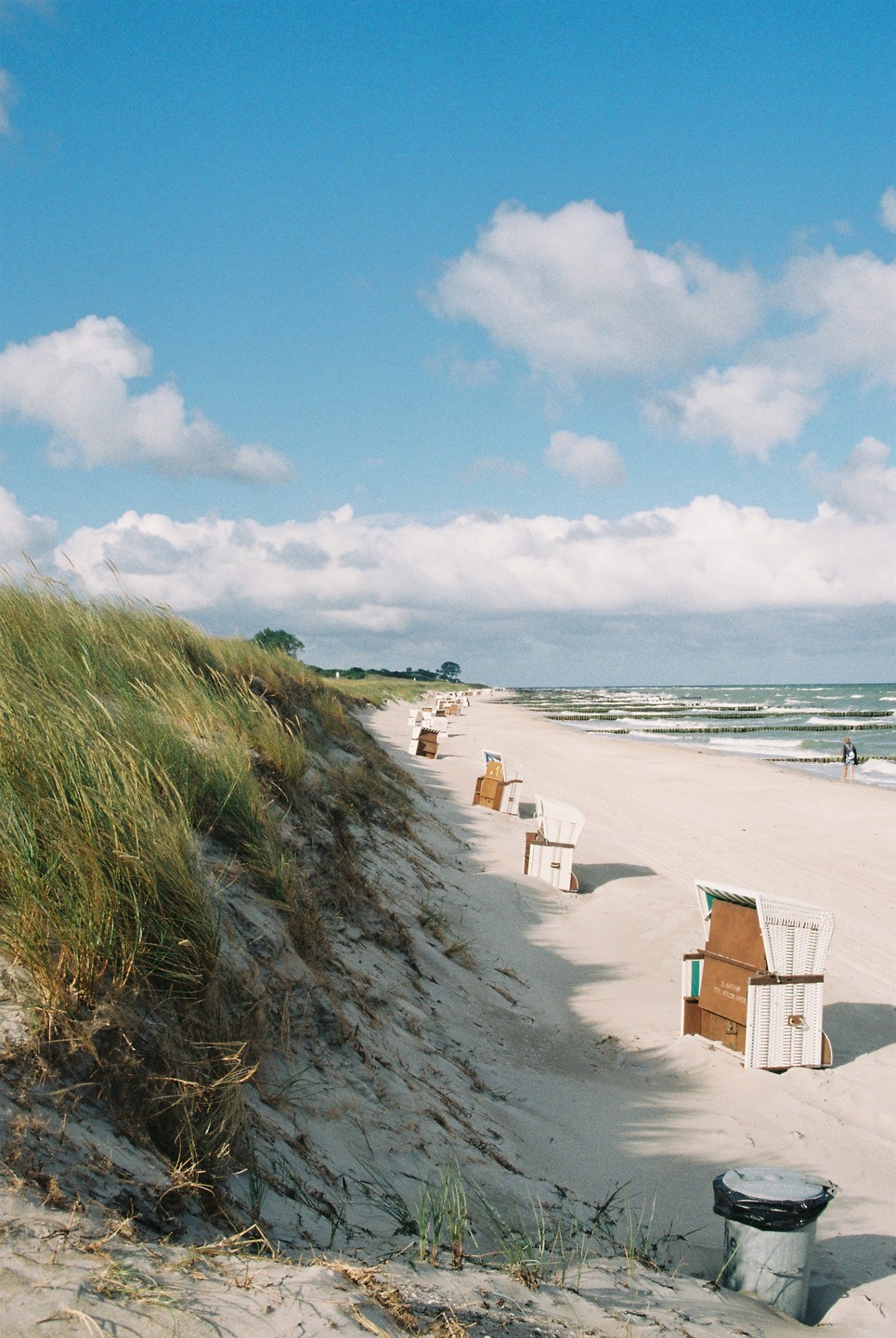 A sandy beach with beach chairs and dunes covered in tall grass stretches along the ocean under a partly cloudy sky.