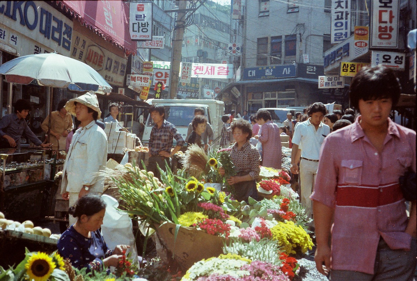 A bustling street market is filled with people, flowers, and various goods under a bright sky.