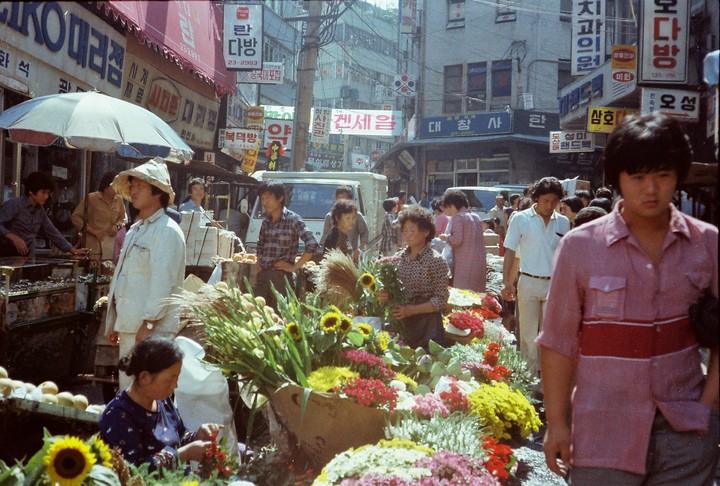 A bustling street market is filled with people, flowers, and various goods under a bright sky.