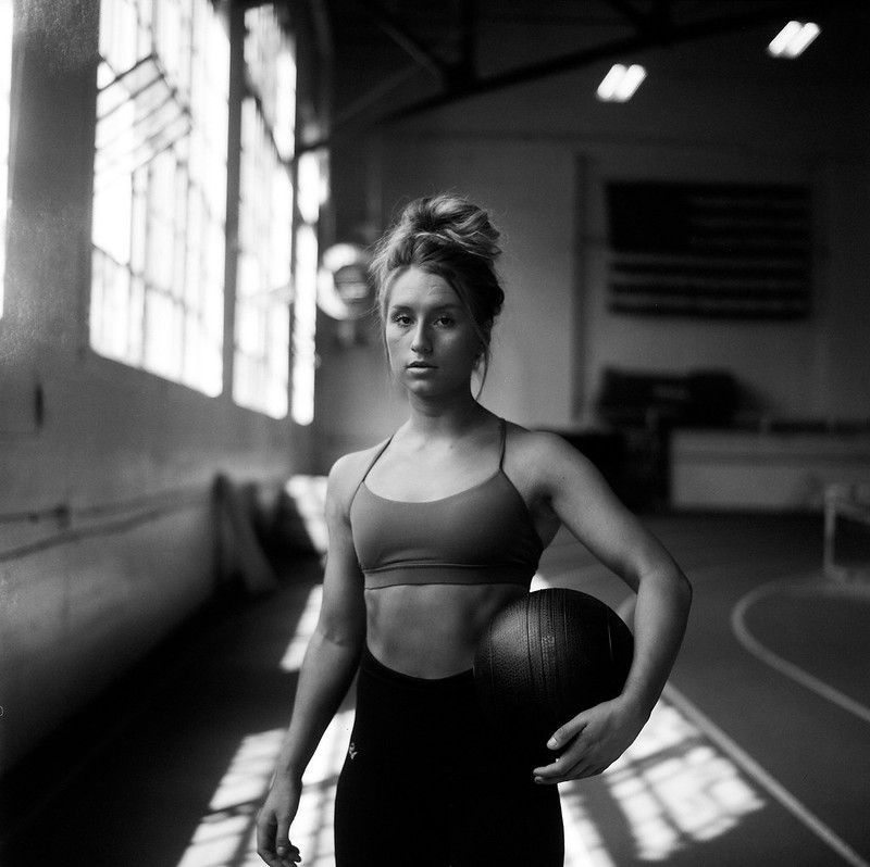 A woman in athletic wear holds a medicine ball in a gym with an American flag on the wall.
