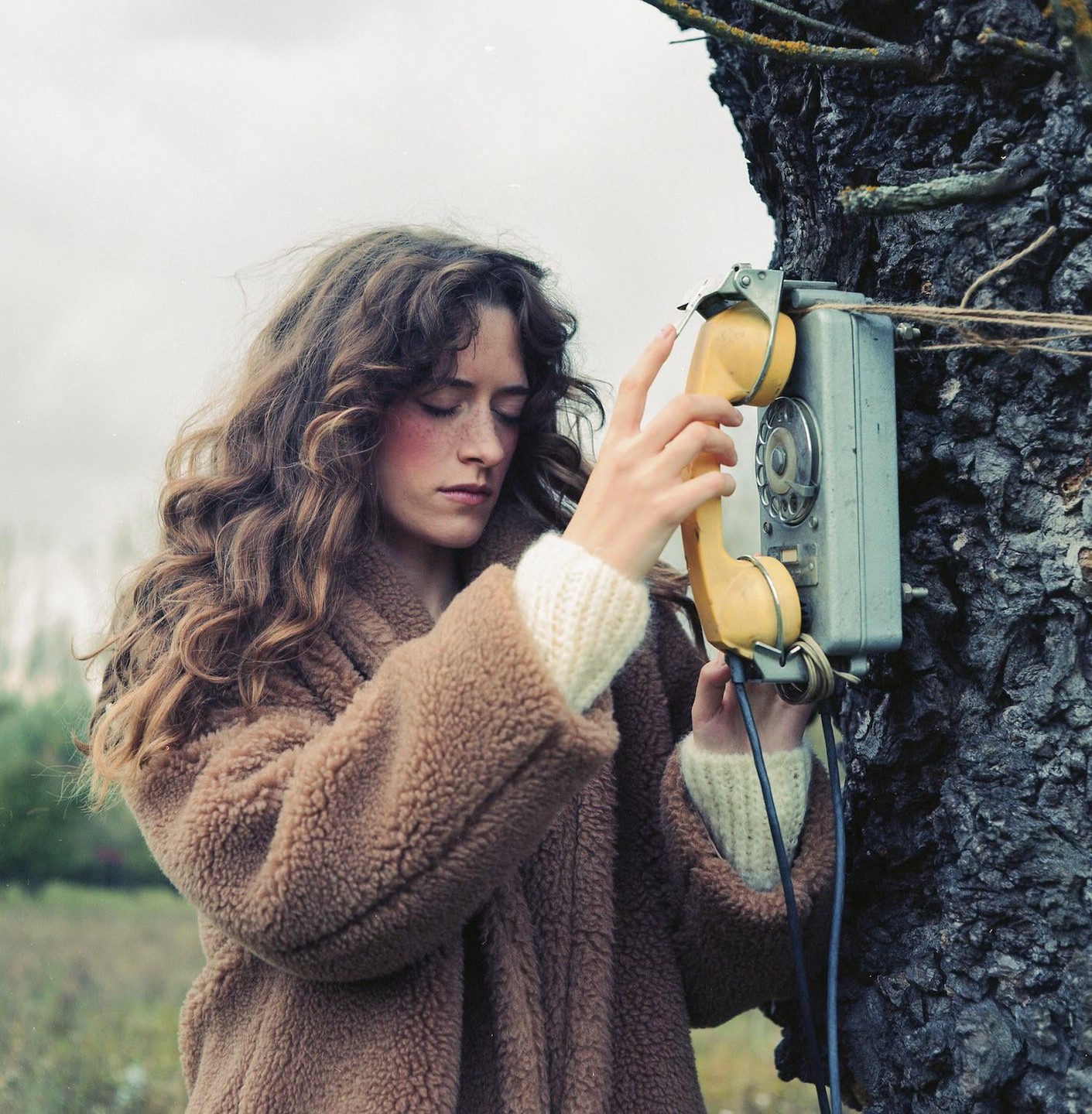 A woman with long, curly hair and freckles holds a yellow rotary phone attached to a tree trunk.