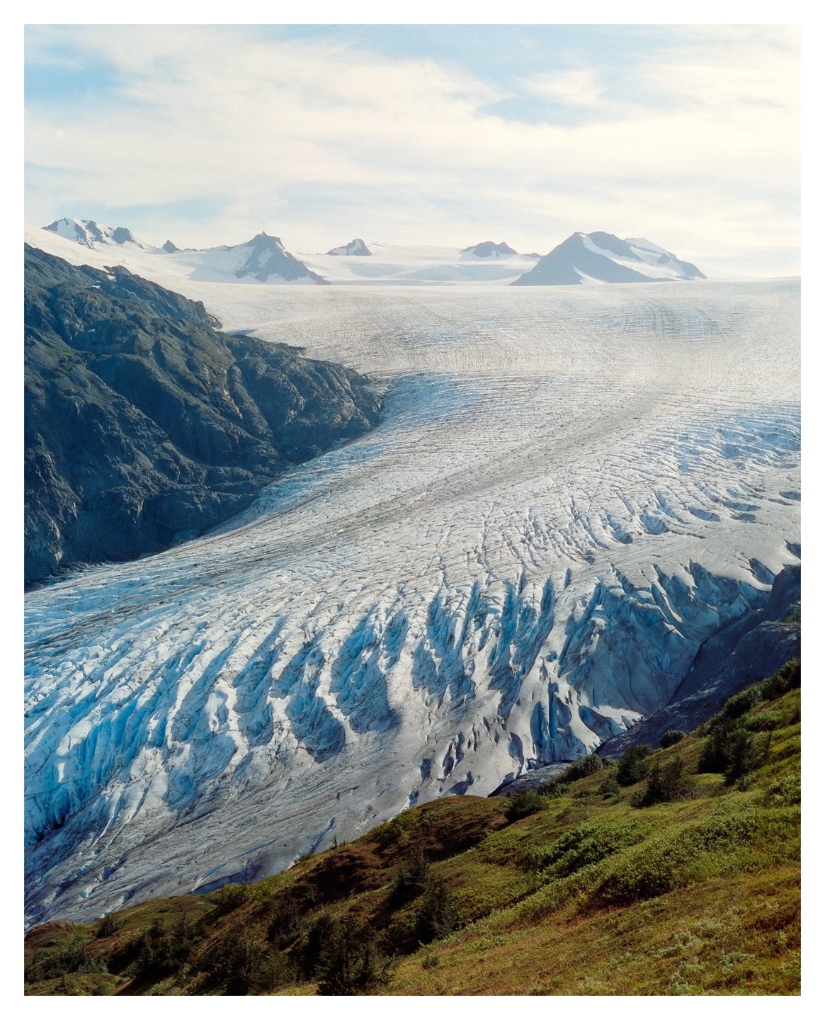 A vast glacier flows between a rocky mountain and a green hillside under a cloudy sky.