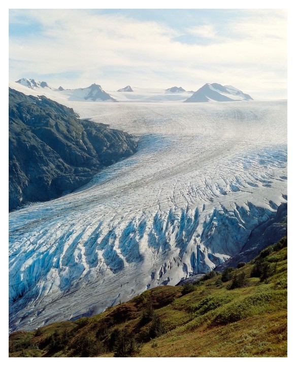 A vast glacier flows between a rocky mountain and a green hillside under a cloudy sky.