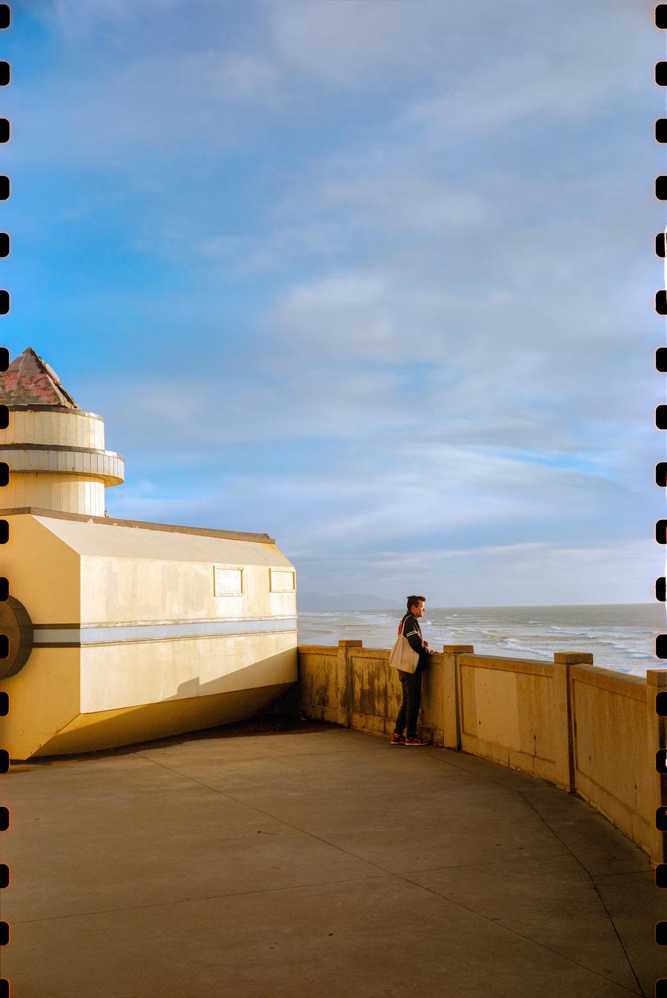 A man stands on a concrete promenade next to a building, looking out at the ocean.
