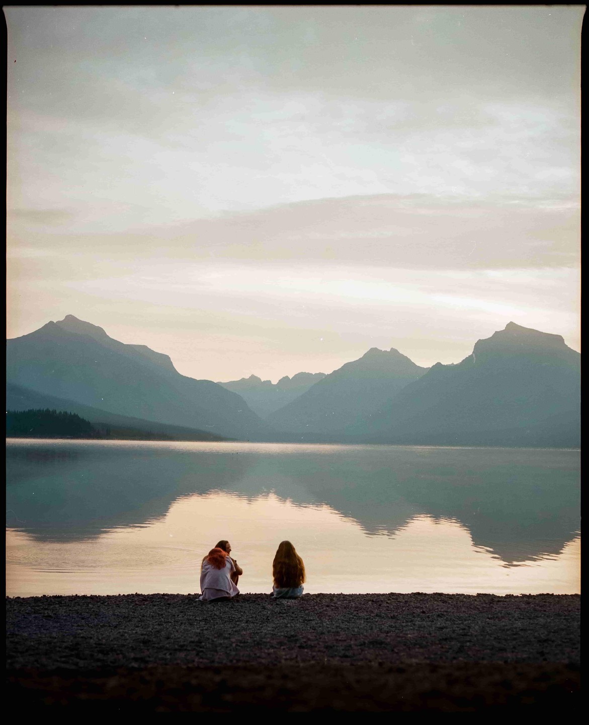 Two people sit on a rocky shore looking out at a lake with mountains in the background under a cloudy sky.