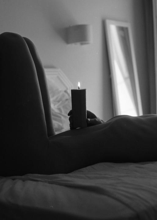 A person lies on a bed holding a lit candle, with a mirror visible in the background.