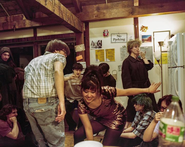 A group of young adults are gathered in a dorm room, some standing and some sitting on the floor, with one woman smoking a cigarette.