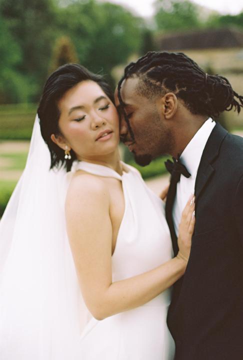 A couple embraces with their eyes closed, the woman wearing a white dress and the man wearing a tuxedo.