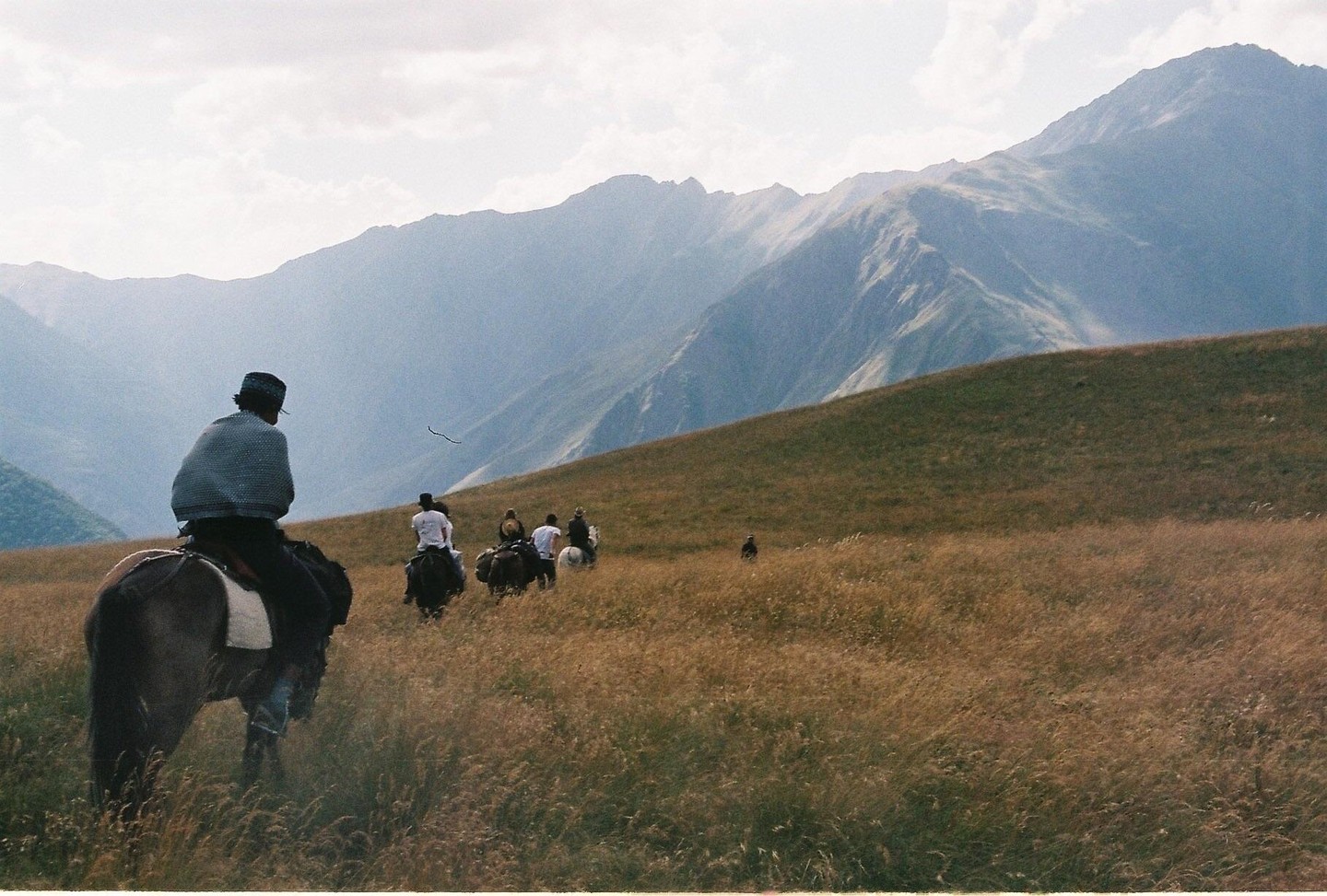 A group of people ride horses through a vast, grassy mountain landscape under a cloudy sky.