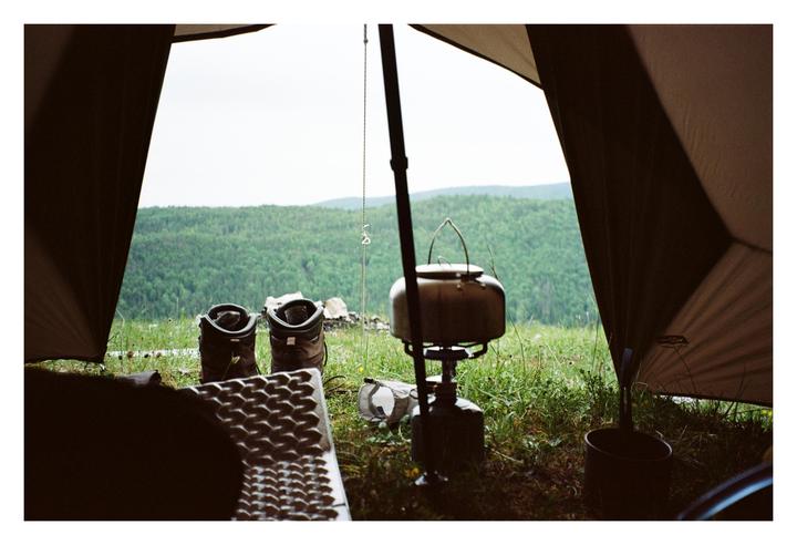 A camping tent opens to a view of a forest and mountains, with hiking boots and a kettle on a stove in the foreground.