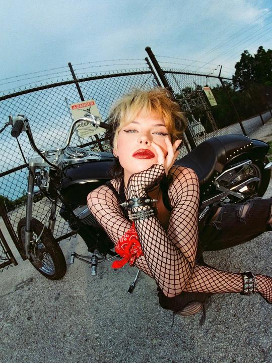 A woman with red lipstick and fishnet clothing poses next to a motorcycle.