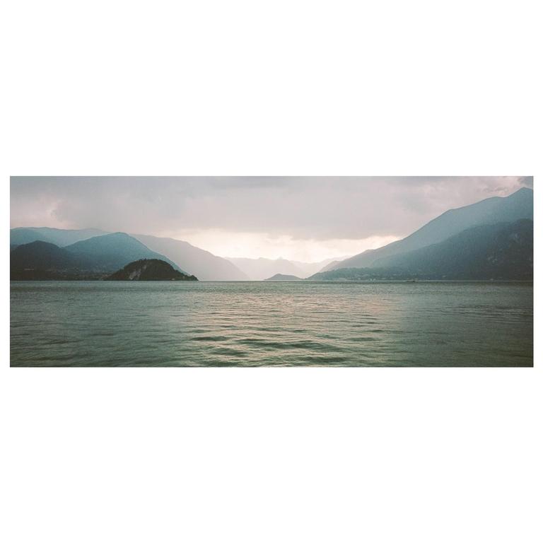 A panoramic landscape shows a lake surrounded by mountains under a cloudy sky.