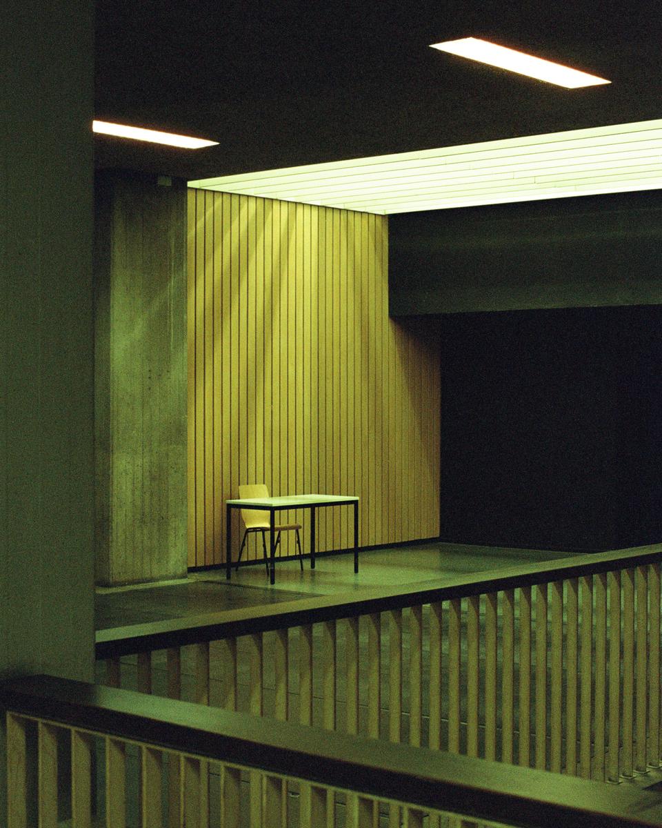 A table and two chairs sit in a room with wood paneling and concrete walls, viewed from above a railing.