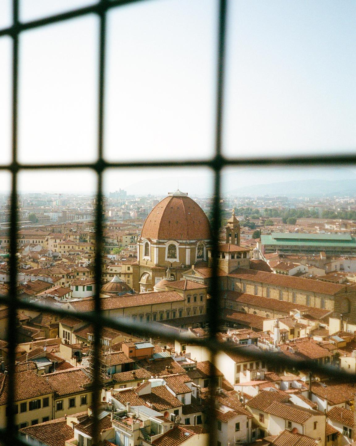 A grid of bars frames a cityscape with a large domed building and many terracotta roofs under a hazy sky.