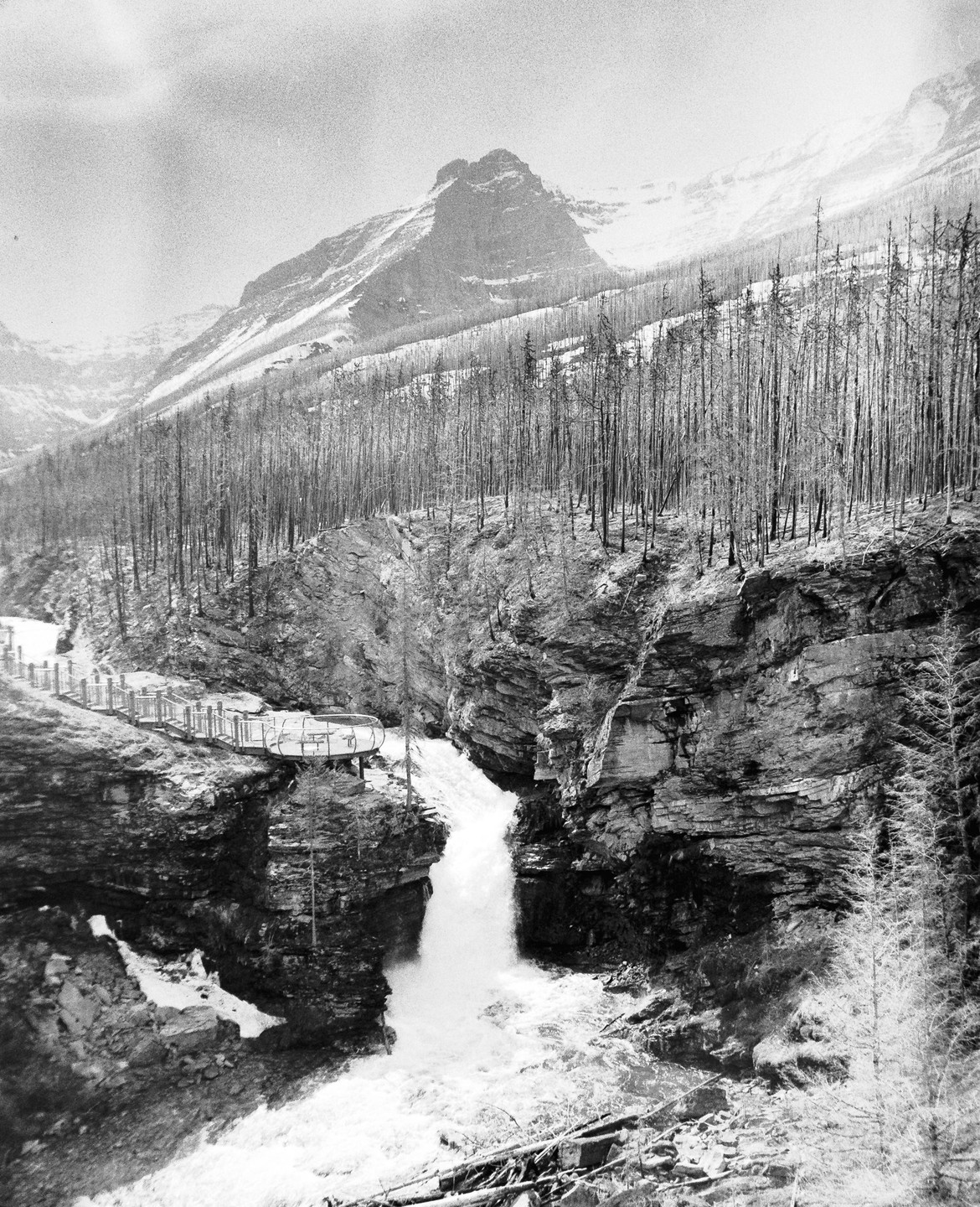 A waterfall cascades through a rocky canyon with a viewing platform and a mountain in the background.