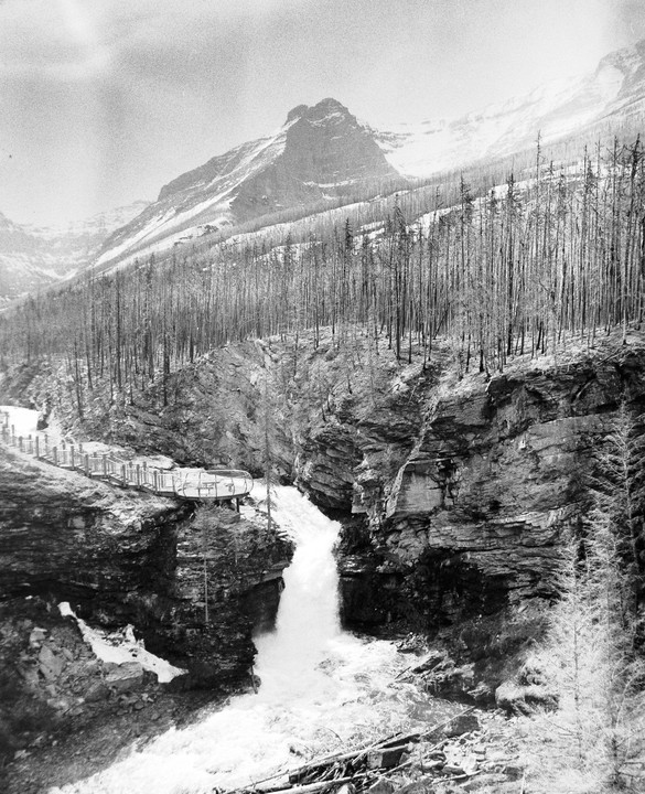 A waterfall cascades through a rocky canyon with a viewing platform and a mountain in the background.