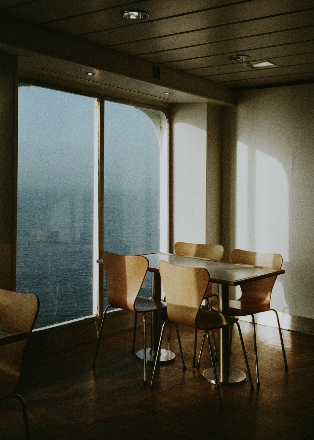 Sunlight streams into an empty dining area on a ferry, illuminating tables and chairs with the sea visible through large windows.