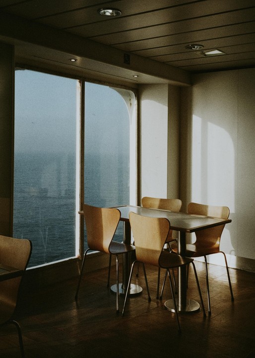 Sunlight streams into an empty dining area on a ferry, illuminating tables and chairs with the sea visible through large windows.