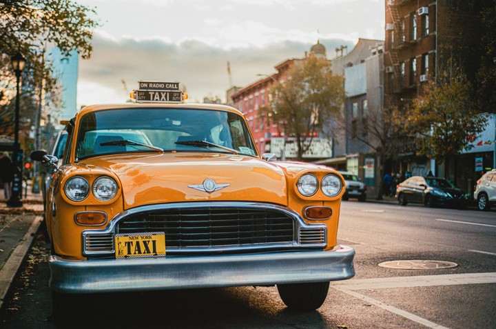 A yellow taxi is parked on a city street with buildings and trees in the background.