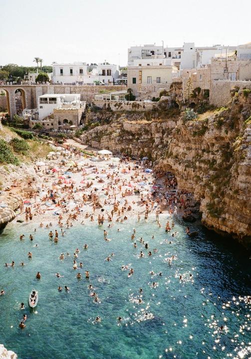 A crowded beach with many people swimming in the clear blue water and sunbathing on the sand, surrounded by cliffs and white buildings.