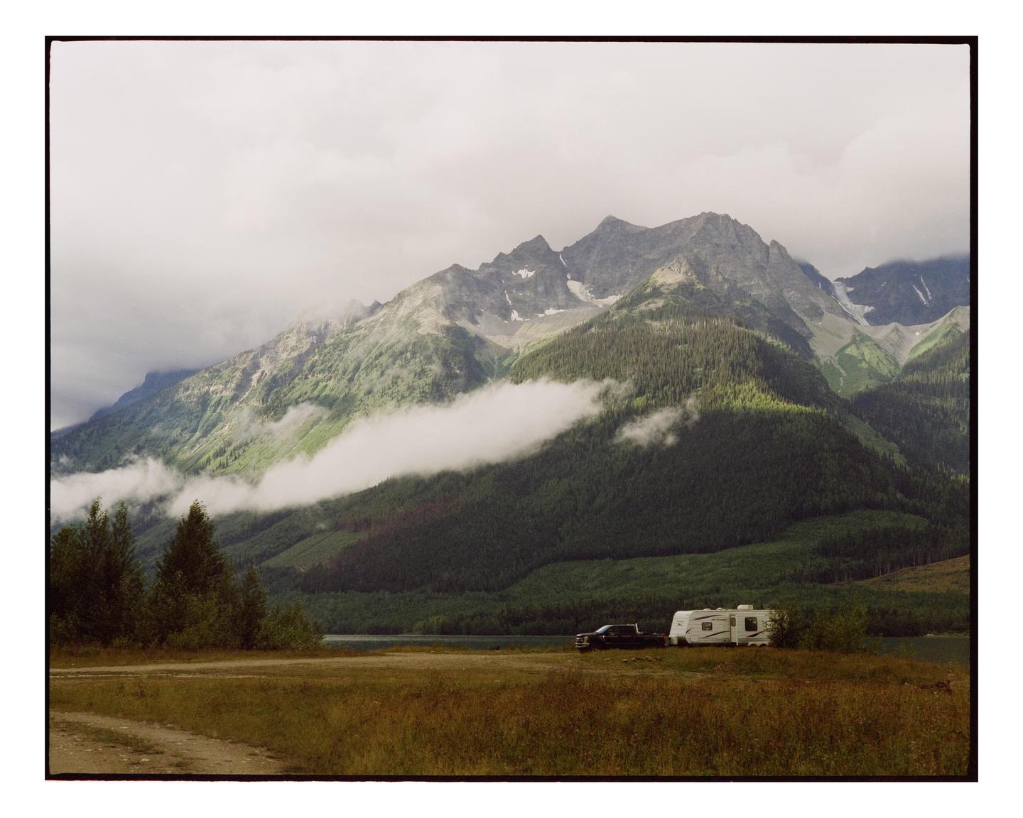 A truck and a camper are parked in a field with a lake and mountains in the background.