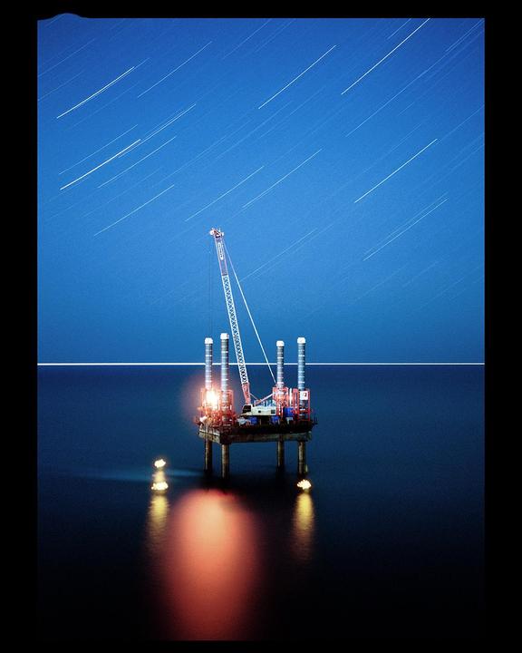 An oil rig with a crane stands in the ocean under a night sky with star trails.