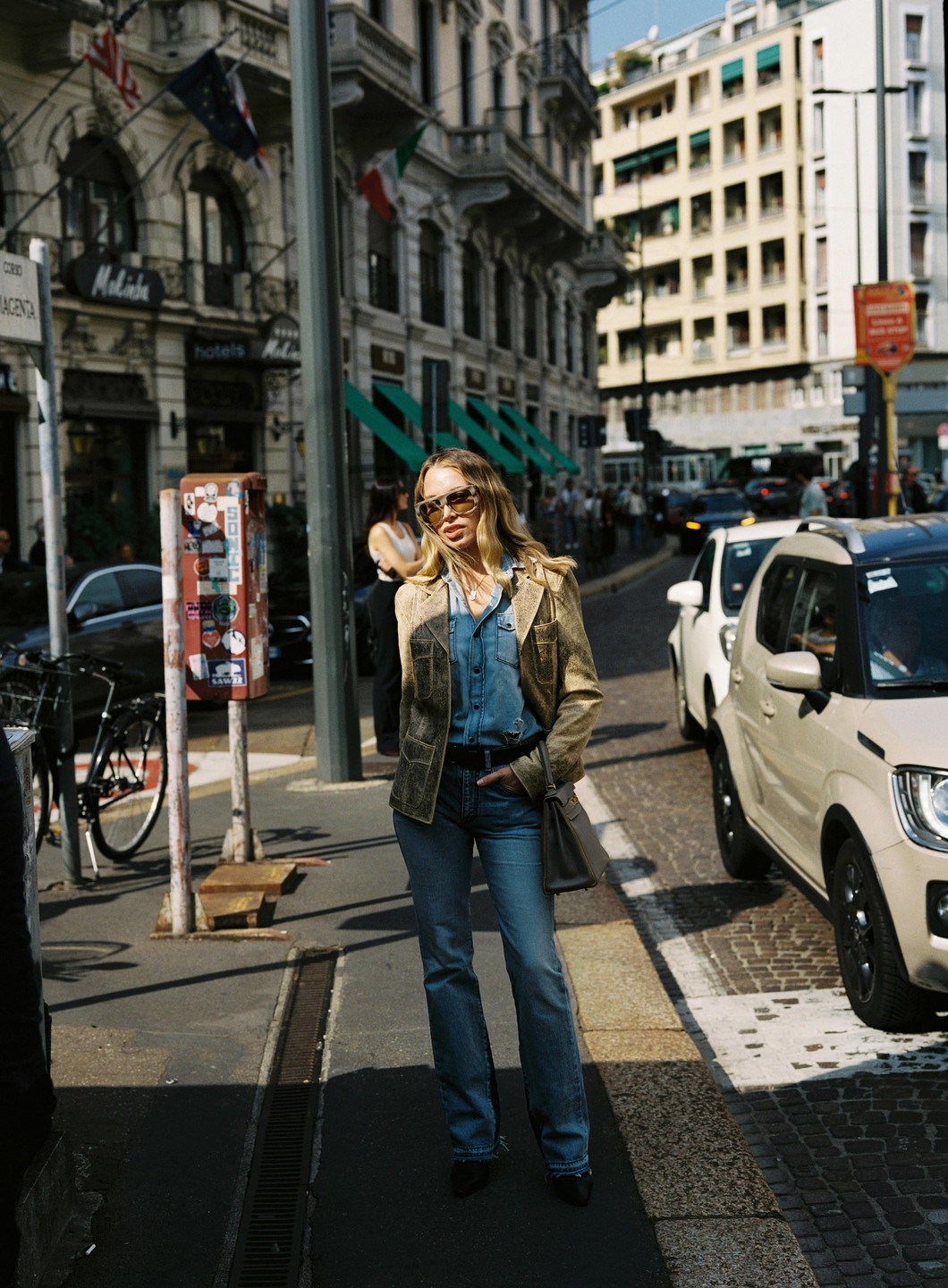 A woman wearing sunglasses, a denim shirt, and a jacket stands on a city street with buildings and cars in the background.