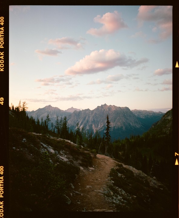 A winding dirt trail leads through a mountainous landscape with pine trees under a sky with pink clouds.