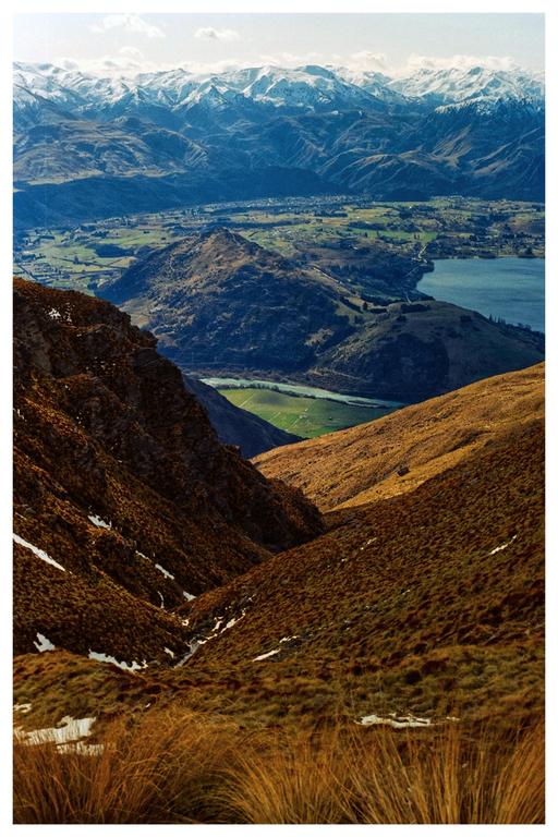 A gully with brown grass and patches of snow leads to a valley with a lake and snow-capped mountains in the distance.