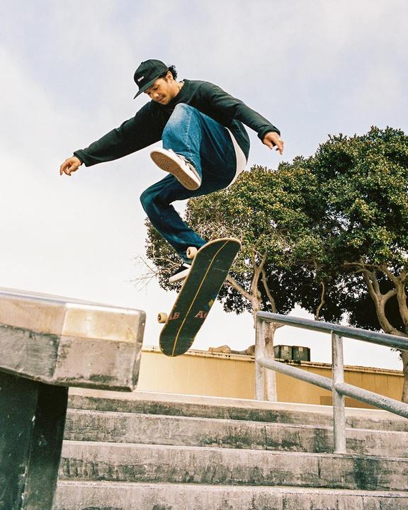 A man in a black sweatshirt and jeans jumps on a skateboard over a concrete railing and stairs.