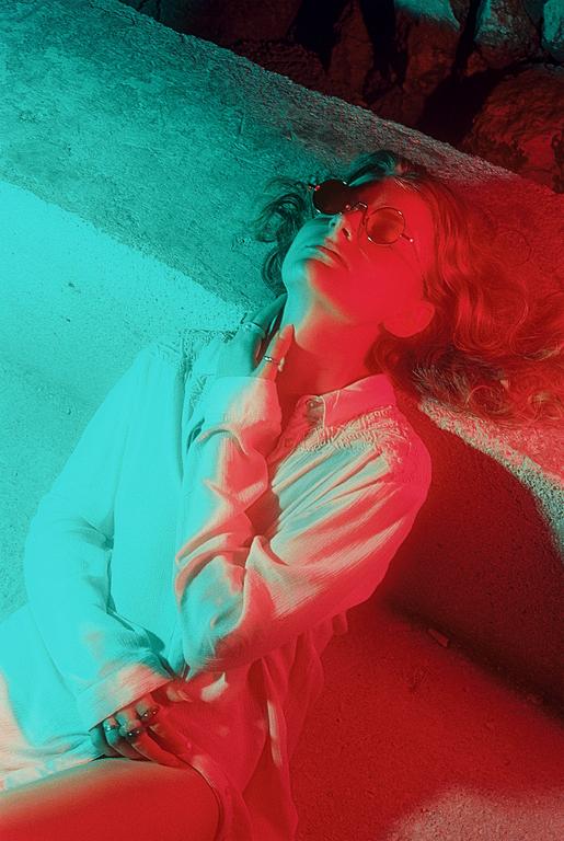 A woman with sunglasses lies on a concrete surface, illuminated by red and blue light.