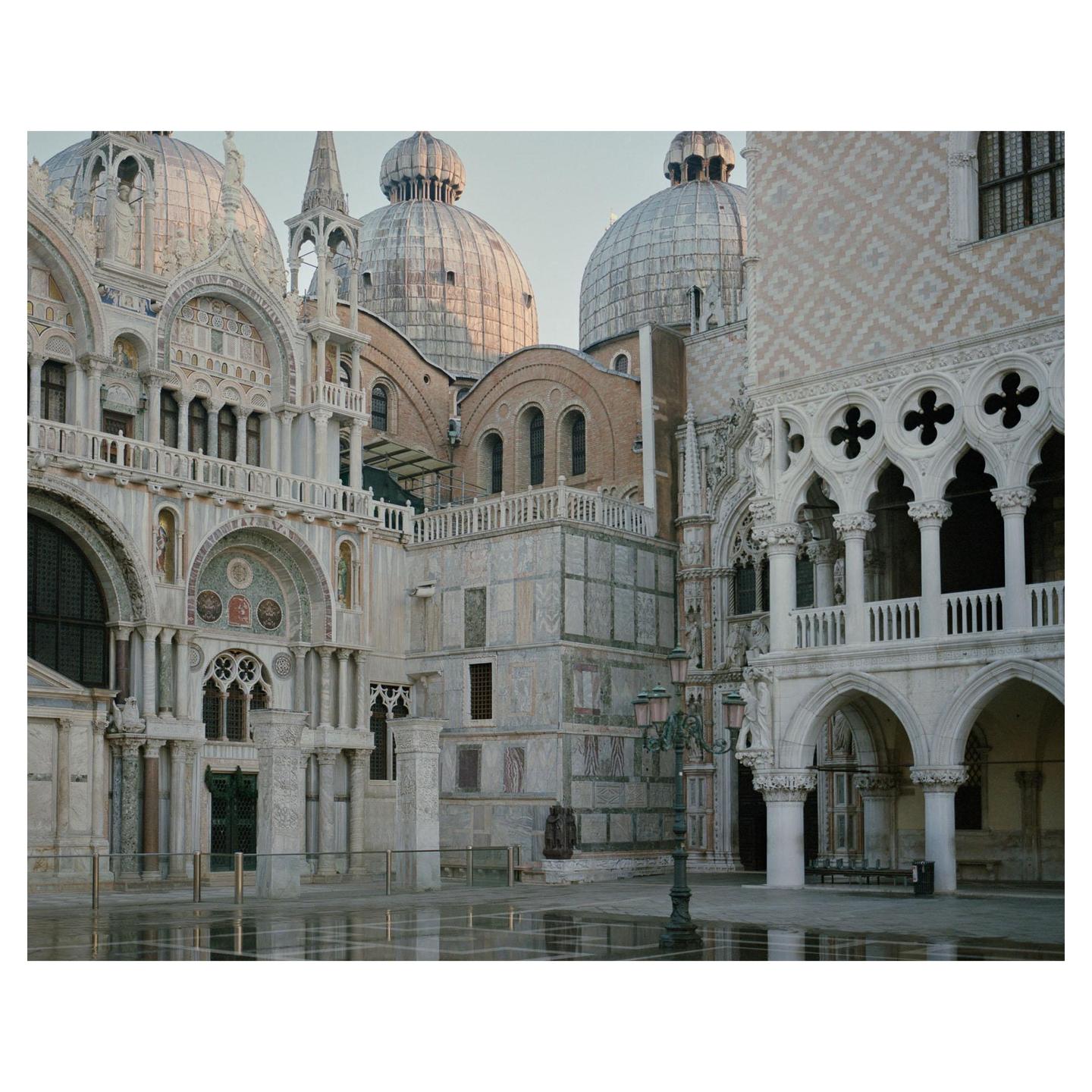 A large ornate building with multiple domes and arches stands under a clear sky, with reflections on the wet ground in the foreground.