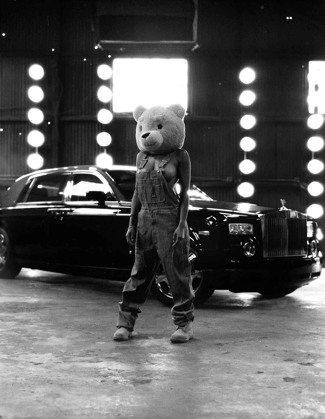 A person wearing a teddy bear head and overalls stands in front of a luxury car in a garage.