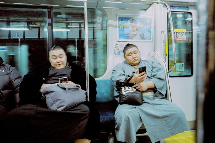 Two sumo wrestlers are sitting on a train, looking at their phones.