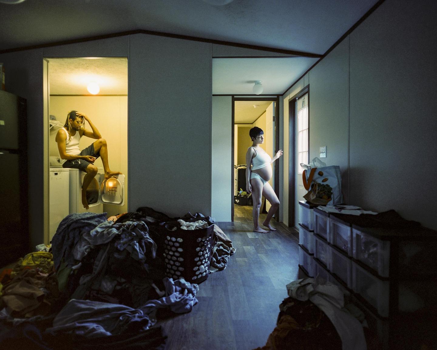 A man sits on a washing machine while a pregnant woman stands in a hallway looking out a door.