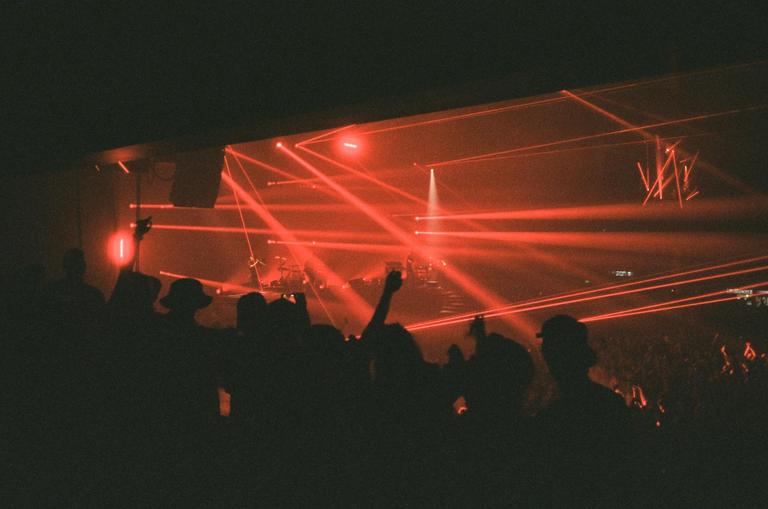 A crowd of people are silhouetted against a stage illuminated by red laser lights at a concert.
