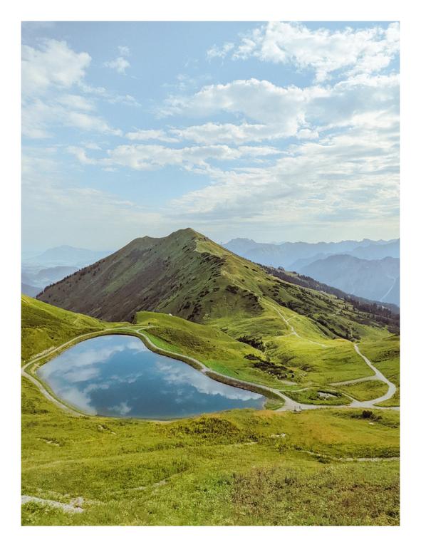 A mountain lake reflects the sky and clouds, surrounded by green hills and mountains under a cloudy sky.