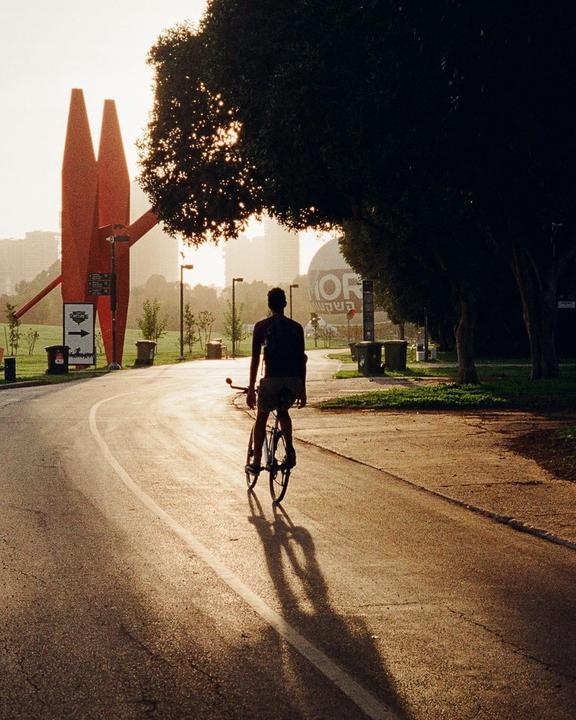 A man rides a bicycle on a paved path with a large red sculpture and trees in the background.