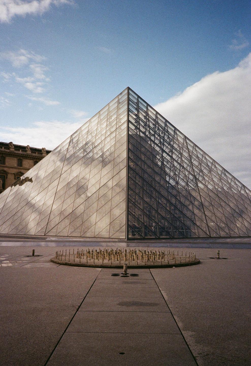 A glass pyramid stands under a blue sky with clouds, with a circular fountain in the foreground.