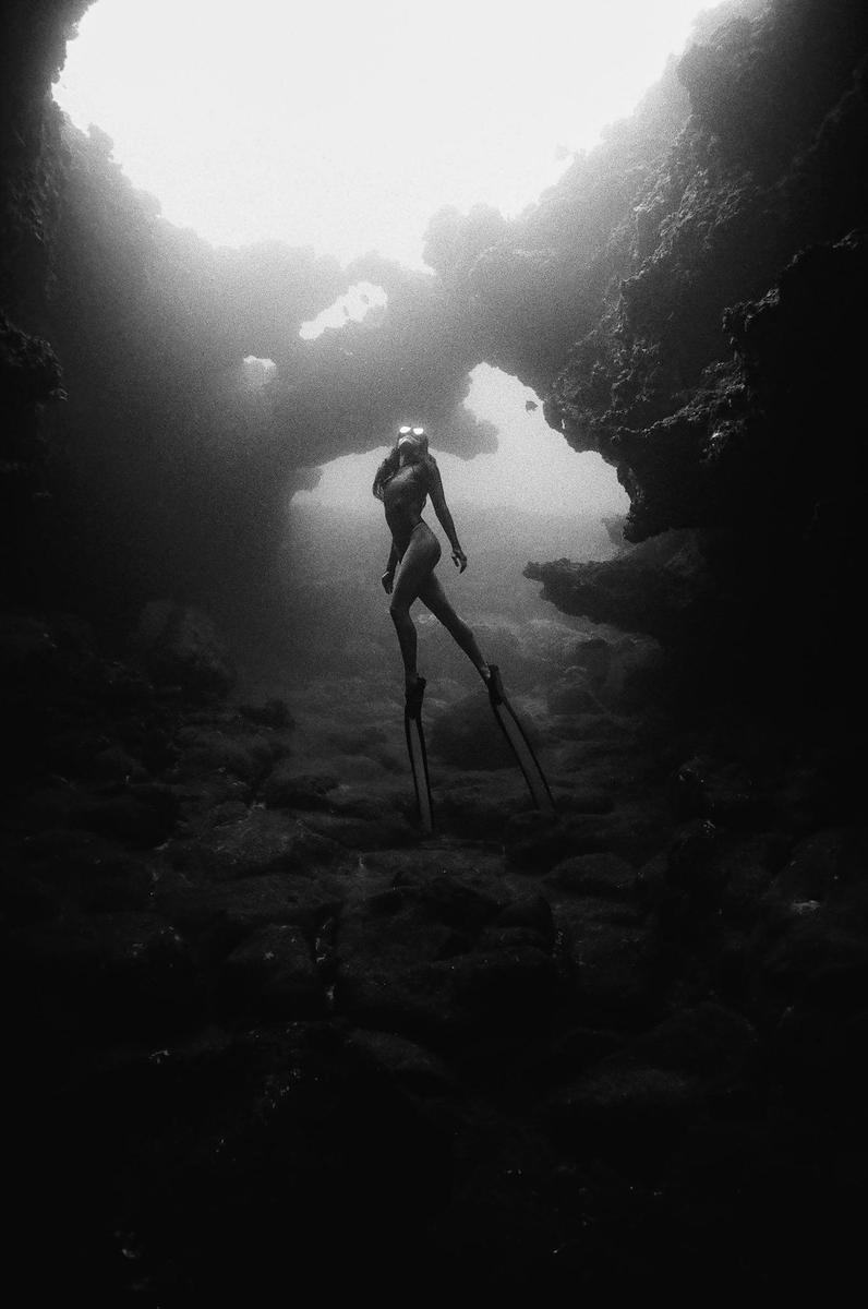 A woman in a swimsuit and flippers is underwater in a dark cave with light coming from above.